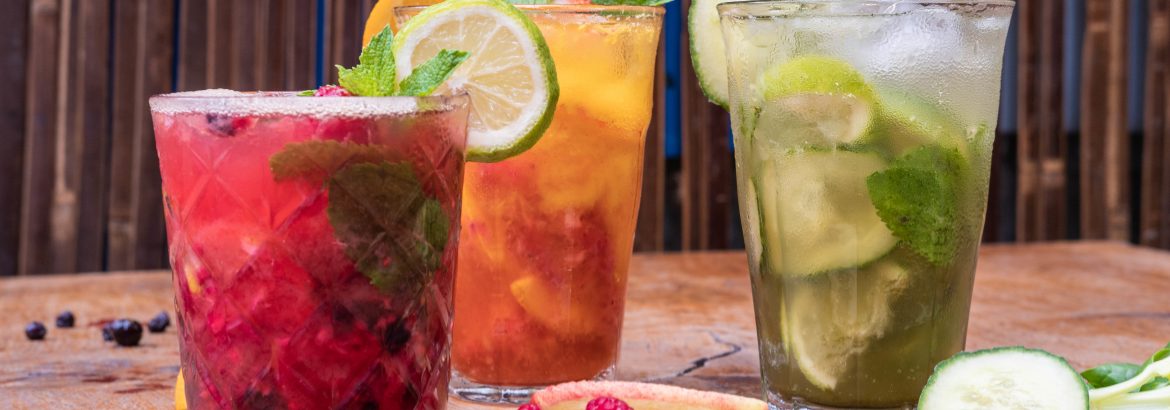 Front view of 3 fresh fruit juices in glasses on a wooden table, there are cucumber and peach slices on the table, with raspberries and blueberries, glasses garnished with mint leaves, lime and orange slices.